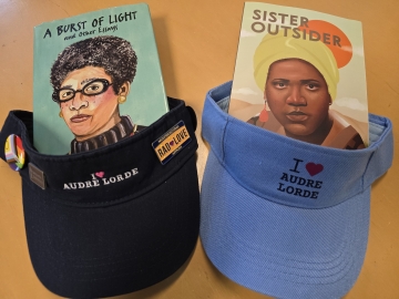 Light blue and Navy blue visors with 'I love Audre Lorde' on them. One has a 'Rad Love' lapel pin on it. Behind the visors are two of Lorde's books with illustrations of her face on the covers..